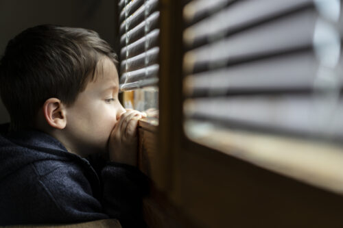 Small sad boy looking through the window experiencing abandonment