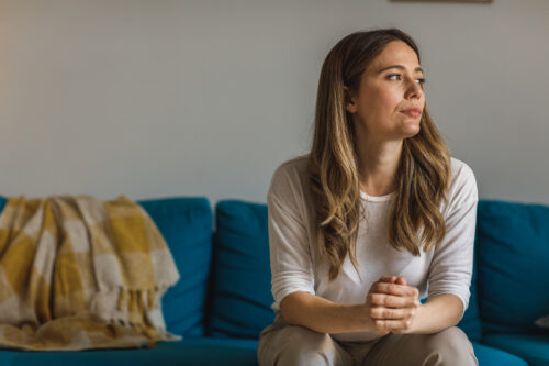 Young woman sitting on the sofa in her living room, cracking her knuckles while contemplating her recent actions and wondering if they're signs of BPD or CPTSD.