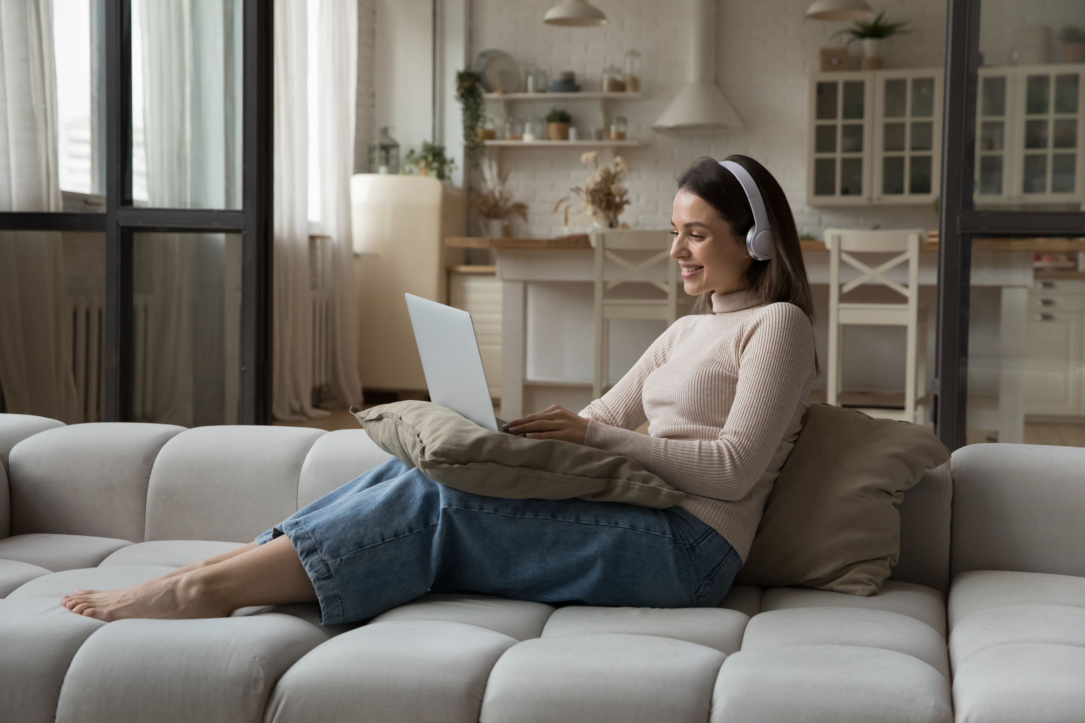 A smiling young woman in headphones using a laptop seated on a cozy sofa attending virtual therapy.