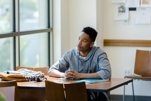 The university student clasps his hands and daydreams while waiting for his study partner in the campus library.